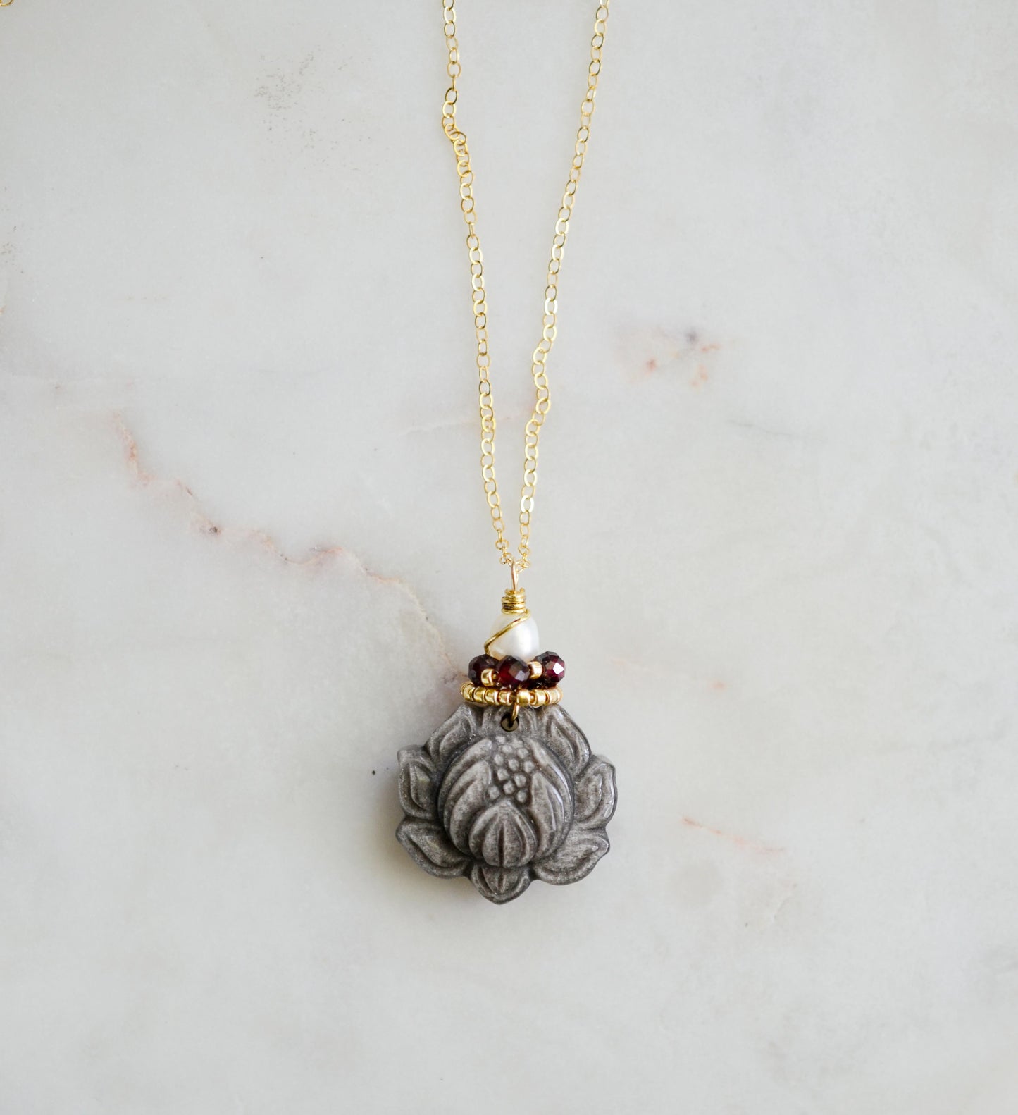 A handmade gemstone pendant featuring a carved obsidian lotus. Garnet, pearl, and gold beads decorate its top. The chain is 14k gold filled.