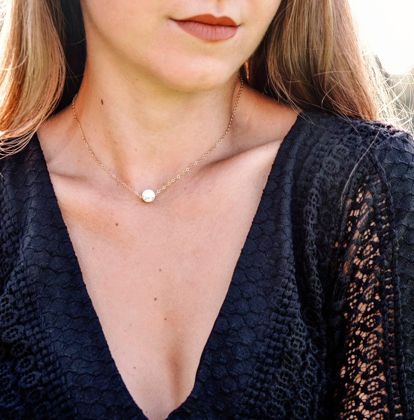 A woman is wearing a natural white howlite necklace. The stone is round with natural gray marbling and set on a 14k gold filled chain. Also available in sterling silver.