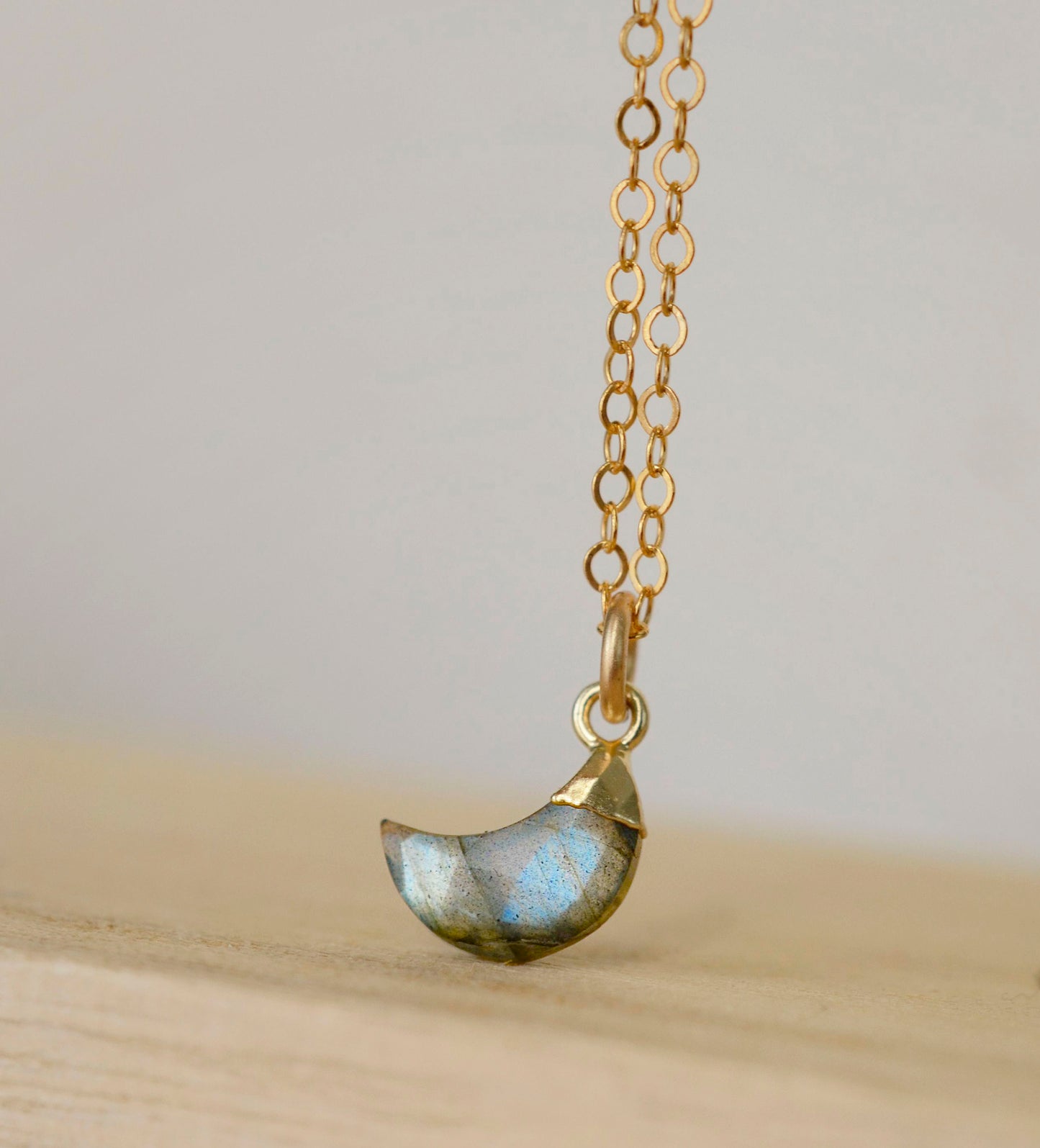 A close up image of a natural labradorite gemstone shaped in a crescent moon and set on a 14k gold filled chain. 