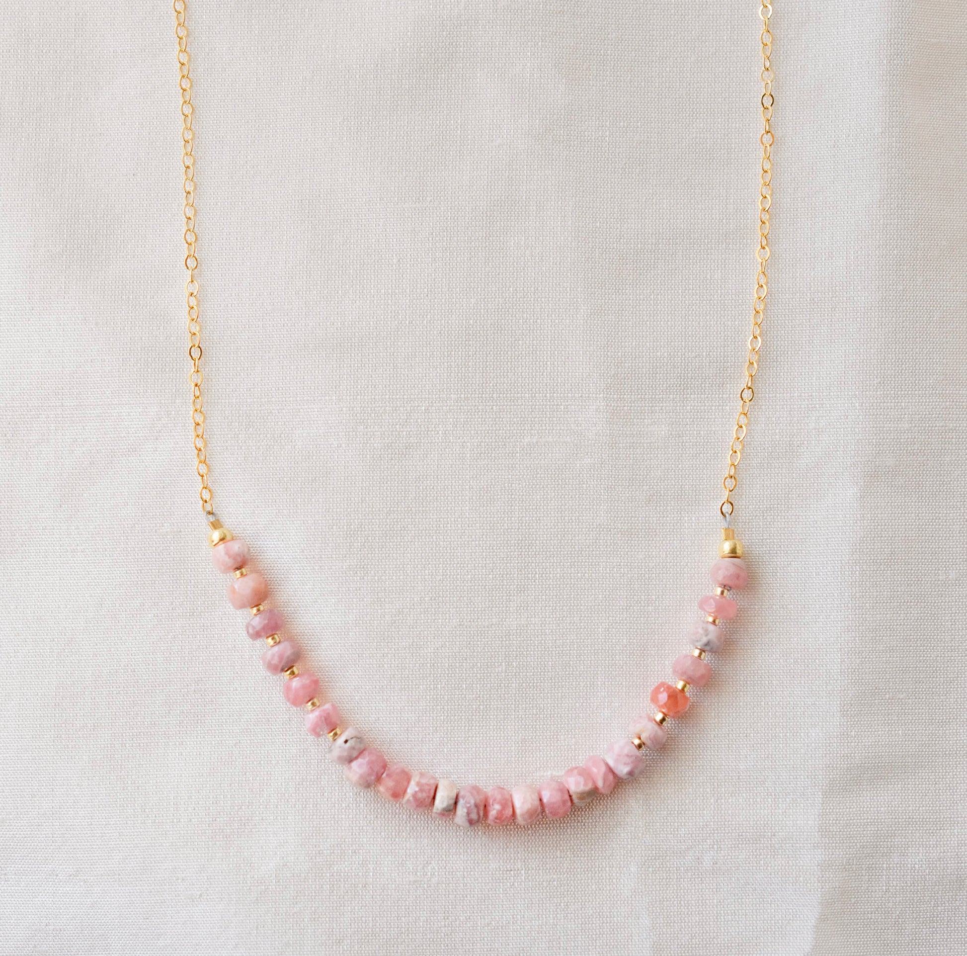 Natural pink Rhodochrosite beaded handmade necklace shown in 14k gold filled. The gemstones vary in shades of pink, peach, white, and gray and are faceted rondelle shape. They span approximately 4 inches across the front of the necklace, then attach to a dainty cable chain. Also available in sterling silver.
