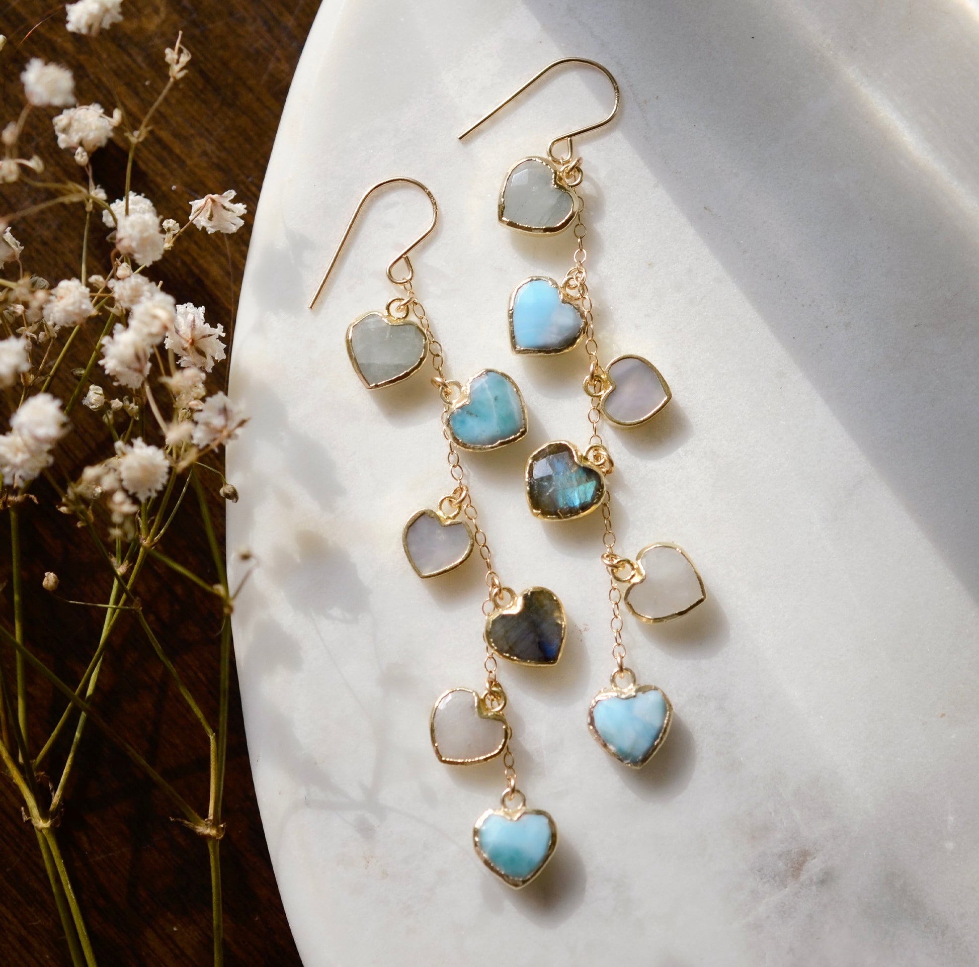 These handmade earrings feature blue and white natural stone hearts bezeled in gold. Each gemstone is a different color and includes: labradorite, larimar, mother of pearl, aquamarine, and rainbow moonstone. Long and light weight they make the perfect romantic gift. They are 3 inches long.