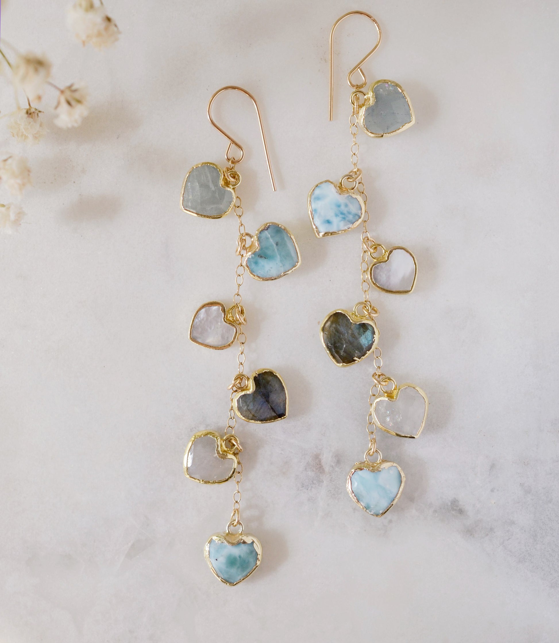 These handmade earrings feature blue and white natural stone hearts bezeled in gold. Each gemstone is a different color and includes: labradorite, larimar, mother of pearl, aquamarine, and rainbow moonstone. Long and light weight they make the perfect romantic gift. They are 3 inches long.