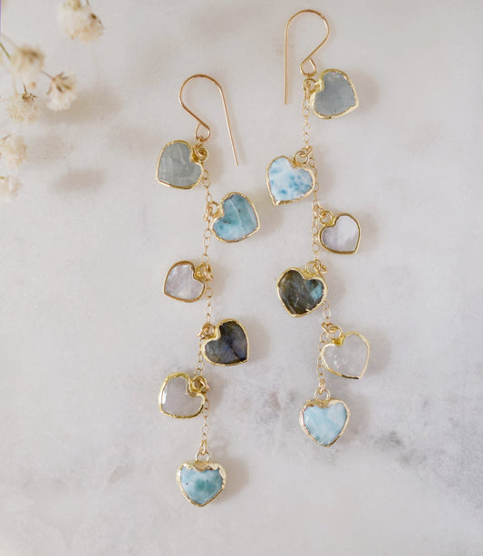 These handmade earrings feature blue and white natural stone hearts bezeled in gold. Each gemstone is a different color and includes: labradorite, larimar, mother of pearl, aquamarine, and rainbow moonstone. Long and light weight they make the perfect romantic gift. They are 3 inches long.