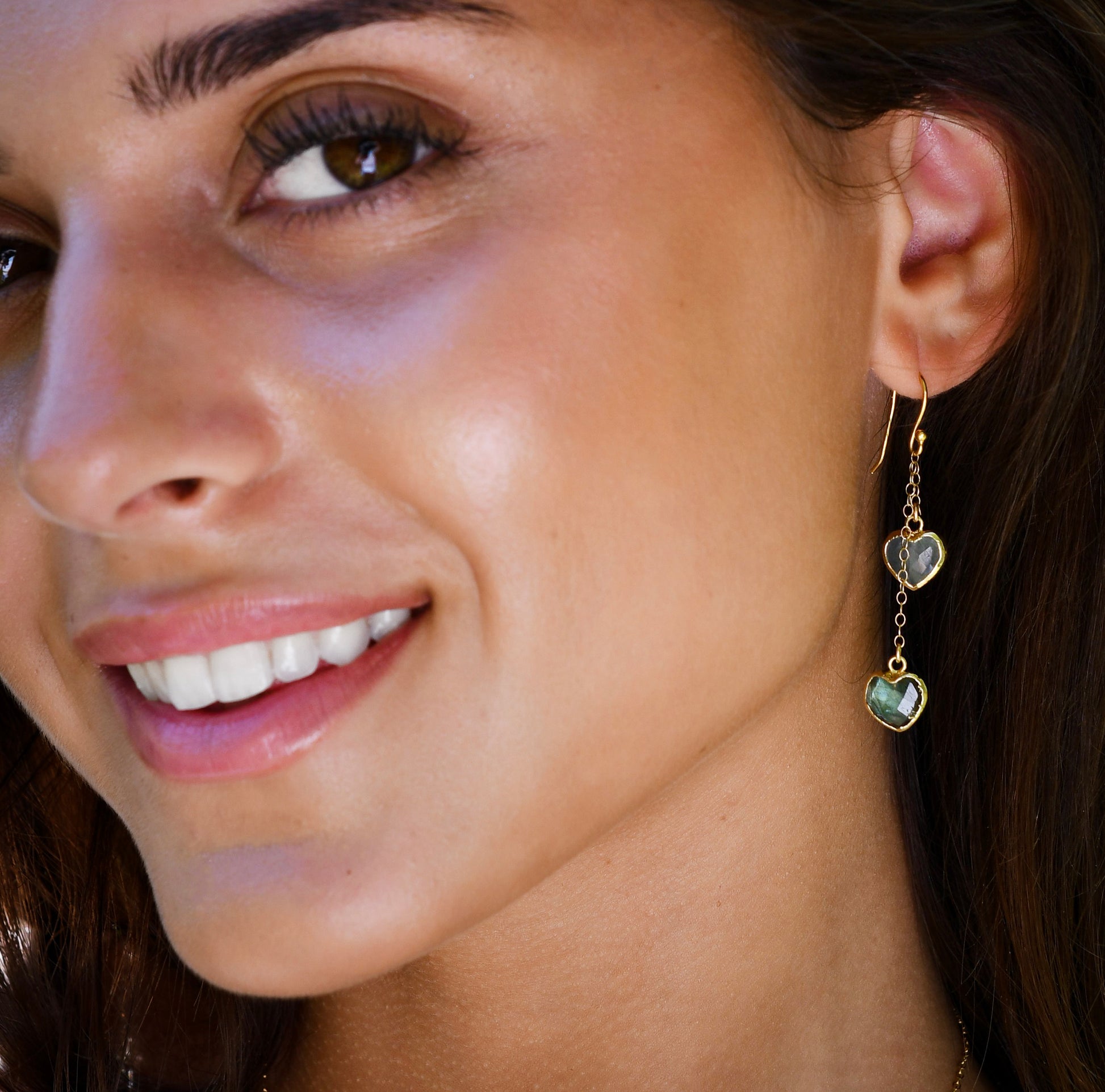 Close-up of a woman with wearing stone heart dangle earrings. The gold gemstone dangle earrings feature aquamarine and labradorite hearts. They're a perfect romantic gift for her, wife, girlfriend, valentines day.