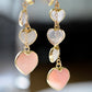 Long pink stone heart earrings in 14k gold filled. There are five heart crystal dangles hanging from a simple chain forming a cluster. The stones include: rose quartz, mother of pearl, pink opal, and rainbow moonstone. They are approximately three inches long.