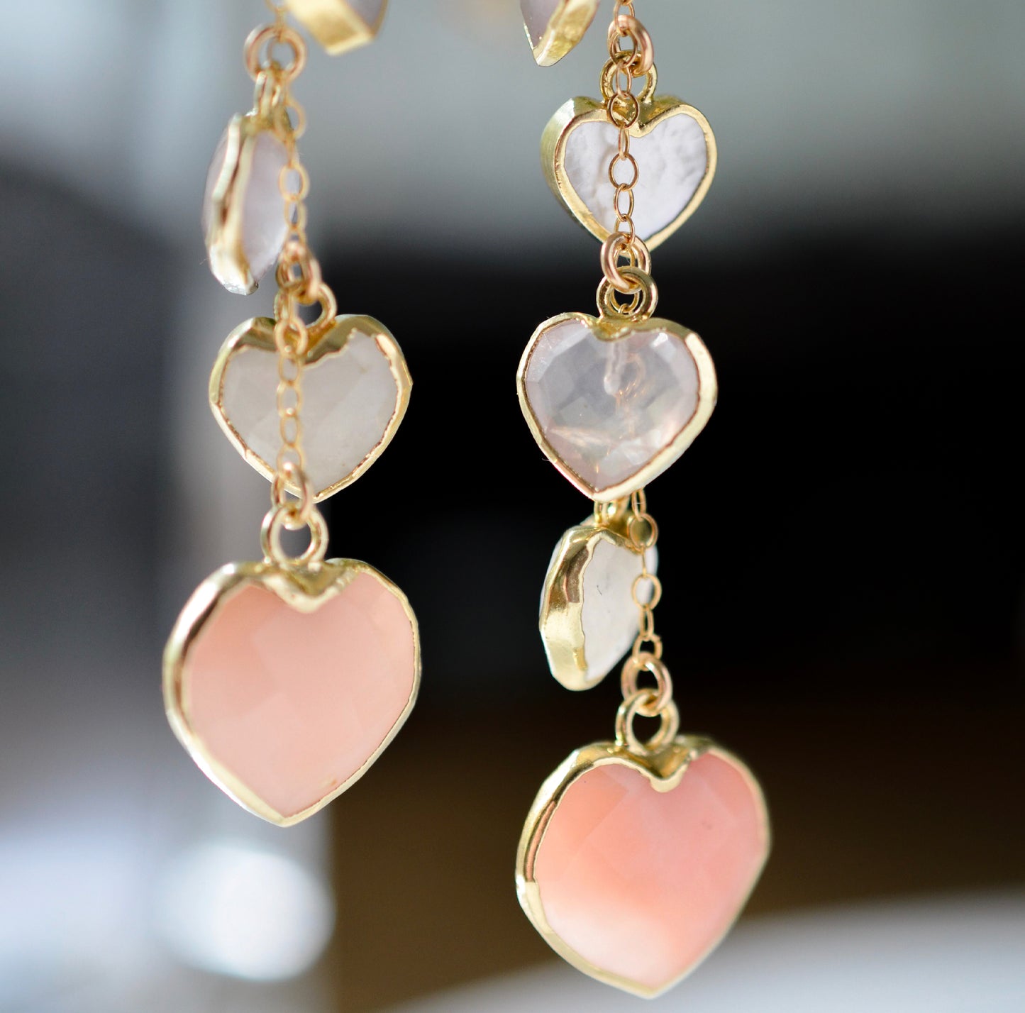 Long pink stone heart earrings in 14k gold filled. There are five heart crystal dangles hanging from a simple chain forming a cluster. The stones include: rose quartz, mother of pearl, pink opal, and rainbow moonstone. They are approximately three inches long.