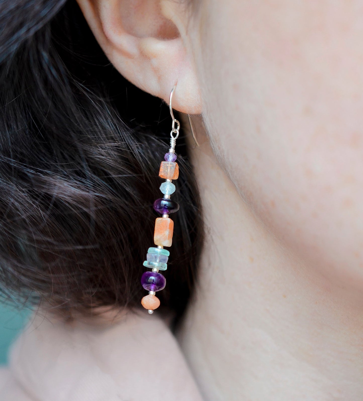 Mismatched gemstone long bar earrings shown in sterling silver. Stones include: amethyst, aquamarine, fluorite, and sunstone. Stones are set onto a long straight bar. Modeled image.