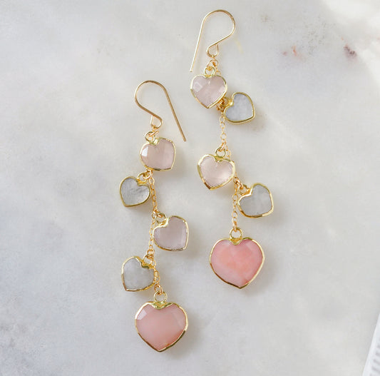 Long pink stone heart earrings in 14k gold filled. There are five heart crystal dangles hanging from a simple chain forming a cluster. The stones include: rose quartz, mother of pearl, pink opal, and rainbow moonstone. They are approximately three inches long.