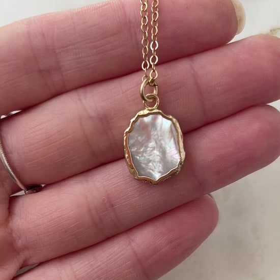 Video. A smooth polished natural, white mother of pearl slice has been set in a 24k gold electroplated bezel and suspended from a 14k gold filled chain. The shell shimmers in shades of white, silver, and pink.