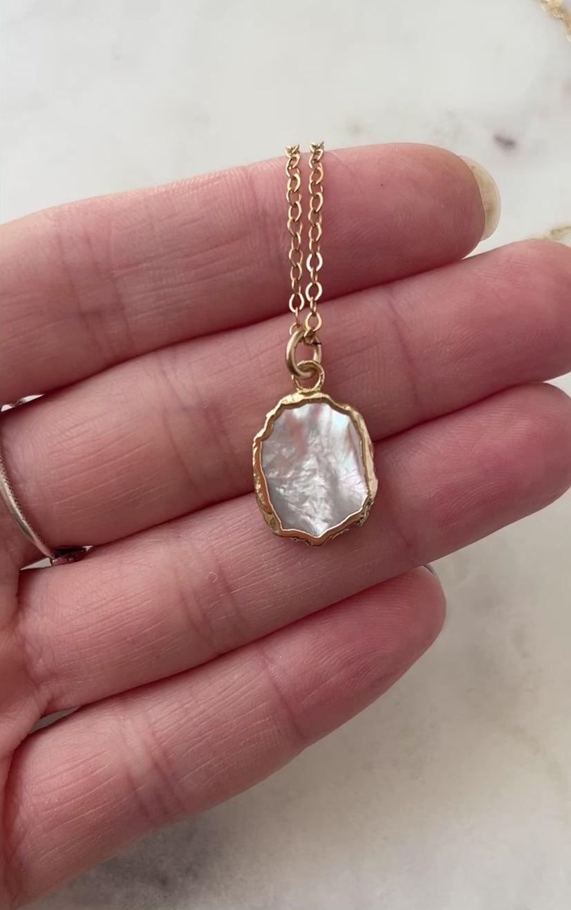 Video. A smooth polished natural, white mother of pearl slice has been set in a 24k gold electroplated bezel and suspended from a 14k gold filled chain. The shell shimmers in shades of white, silver, and pink.