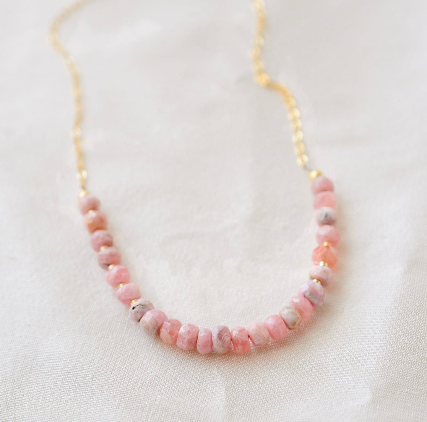 Natural pink Rhodochrosite beaded handmade necklace shown in 14k gold filled. The gemstones vary in shades of pink, peach, white, and gray and are faceted rondelle shape. They span approximately 4 inches across the front of the necklace, then attach to a dainty cable chain. Also available in sterling silver.