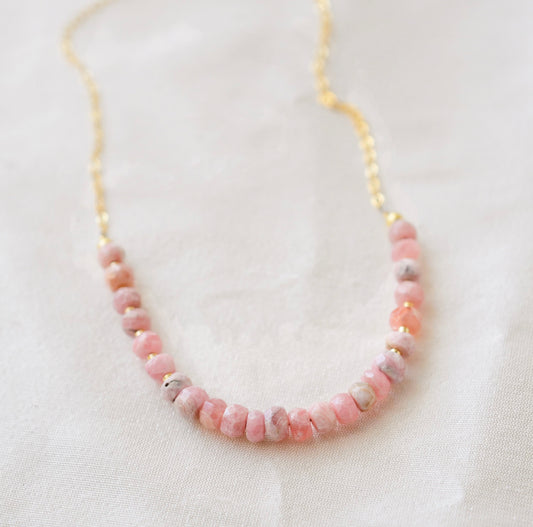 Natural pink Rhodochrosite beaded handmade necklace shown in 14k gold filled. The gemstones vary in shades of pink, peach, white, and gray and are faceted rondelle shape. They span approximately 4 inches across the front of the necklace, then attach to a dainty cable chain. Also available in sterling silver.