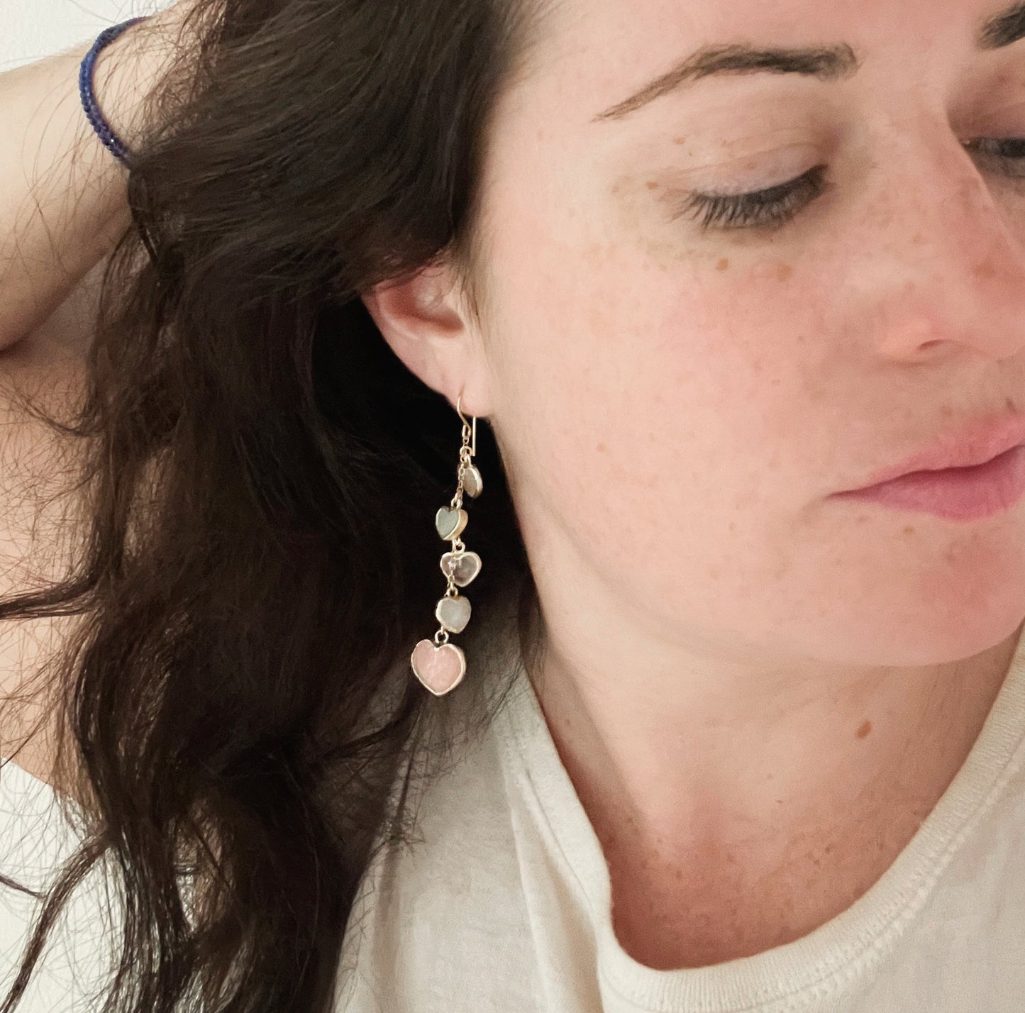 Modeled image. Long pink stone heart earrings in 14k gold filled. There are five heart crystal dangles hanging from a simple chain forming a cluster. The stones include: rose quartz, mother of pearl, pink opal, and rainbow moonstone. They are approximately three inches long.