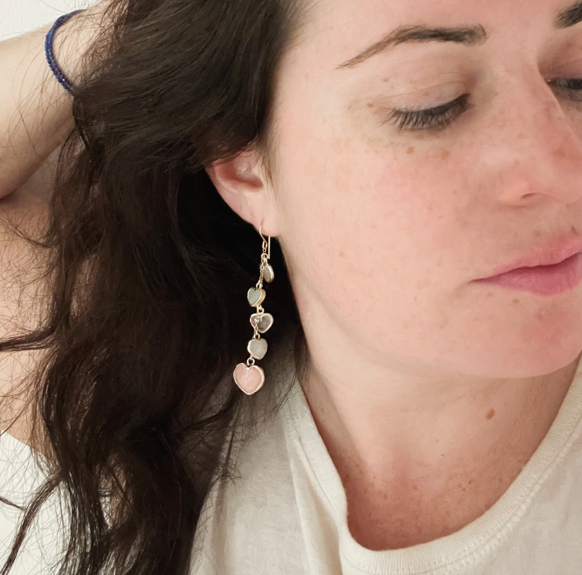 Modeled image. Long pink stone heart earrings in 14k gold filled. There are five heart crystal dangles hanging from a simple chain forming a cluster. The stones include: rose quartz, mother of pearl, pink opal, and rainbow moonstone. They are approximately three inches long.