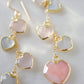 Long pink stone heart earrings in 14k gold filled. There are five heart crystal dangles hanging from a simple chain forming a cluster. The stones include: rose quartz, mother of pearl, pink opal, and rainbow moonstone. They are approximately three inches long.
