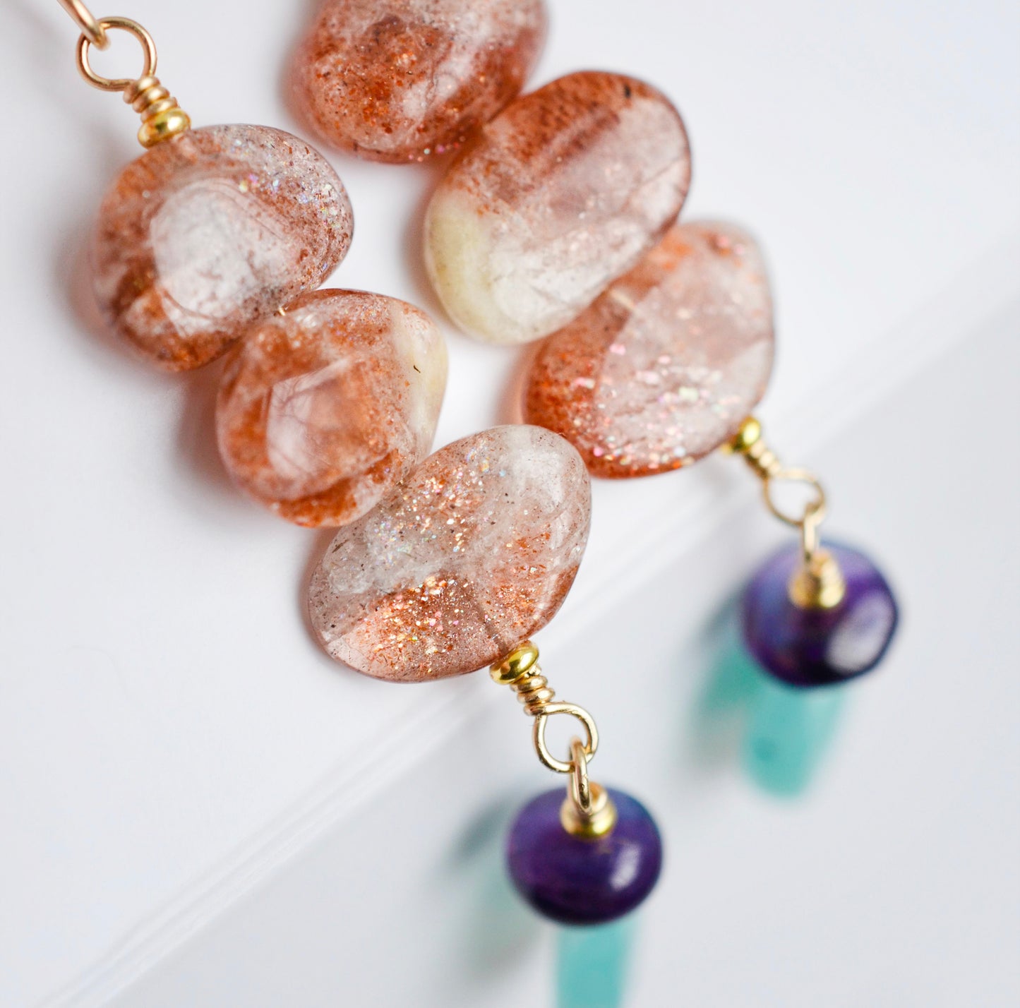 Three sunstone gemstone dangle atop amethyst and amazonite stones. The earring are shown in gold. Close up of the crystals.