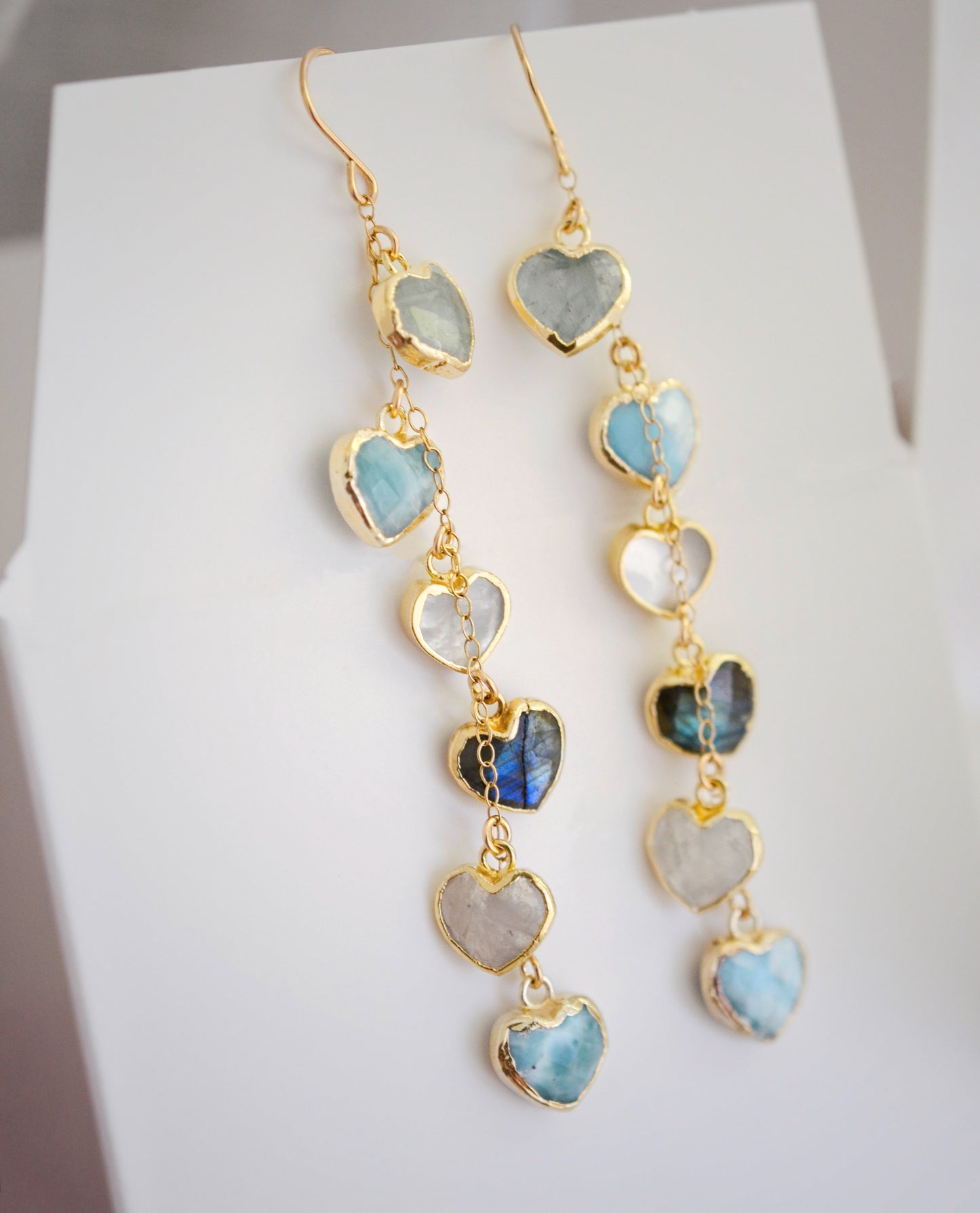 These handmade earrings feature blue and white natural stone hearts bezeled in gold. Each gemstone is a different color and includes: labradorite, larimar, mother of pearl, aquamarine, and rainbow moonstone. Long and light weight they make the perfect romantic gift. They are 3 inches long.