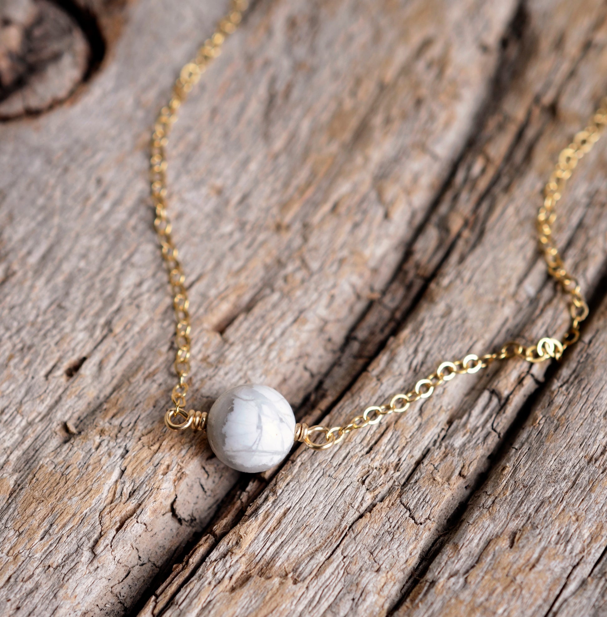 A white howlite necklace on a wood background. The stone is round with natural gray marbling and set on a 14k gold filled chain. Also available in sterling silver. 